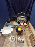 Full lot showing Hamilton Beach slow cooker, various mugs, teapot, measuring cup, egg cups, and CorningWare dishes arranged on wood surface against dark backdrop.