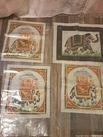 Four unopened hand painted decorative scrolls featuring elephants, three identical and one different, with gold painted embellishments visible through plastic covering on a fabric surface.