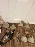 Overview photo showing three electrical wall mount light fixtures on the floor: two double-shade wall sconces with ornate metal backplates and beige frosted glass shades, plus one single lantern-style wall light with dark metal frame and clear glass.