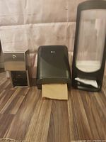 Three dispensers on wood floor background: stainless steel toilet paper dispenser, black plastic paper towel dispenser with brown towels, tall black plastic napkin dispenser.