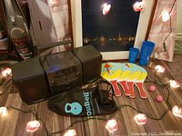 Vintage RCA stereo system with two detachable speakers, Calgary Flames string lights in front, wooden paddles, two pink balls, six blue cups of varying sizes, black backpack, and a faint view of a framed painting in the background.
