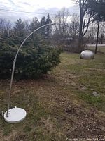 Full view of the tall arc lamp positioned outdoors on grass, showing the white marble base and curved stainless steel arm extending with the dome shade at the end.