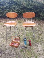 Two wooden kitchen stools with curved seats and metal legs, placed outdoors with jewelry box, thermos, plastic shaker cup, and bamboo tea light holder displayed in front.