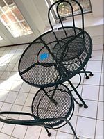 Photo showing green metal mesh round table with two matching mesh chairs indoors on tile floor against windowed door.