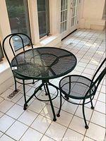 Photo showing the entire round metal mesh table with two matching chairs placed on tiled floor near windows.