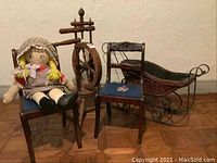Full view of the 5-piece children's decor set showing rattan doll sleigh, wooden spinning wheel, two child's size wooden chairs with embroidered needlepoint seats, and a rag doll sitting on one chair.