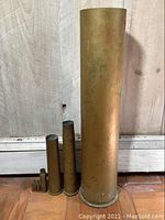 Full view of seven brass artillery shell casings of varying sizes standing upright against a wall on the floor.