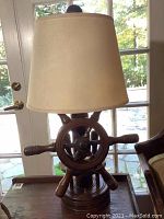 Front and side views of the ship's wheel wooden table lamp with beige shade on a wooden side table.