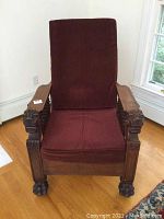 Front view of vintage Morris chair with burgundy cushions and carved wooden arms ending in lion heads as well as claw feet.
