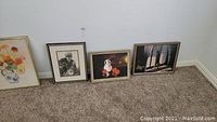 Four framed artworks displayed on carpet leaning against wall with white baseboard.