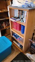 Front view of a wood veneer bookcase filled with books and some items on top, showing three shelves and medium brown wood grain surface.