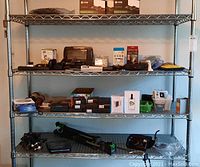 Wide view of shelving holding electronic devices, clocks, tape recorder, watches, batteries, and accessories.