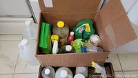 Top view of two boxes filled with assorted cleaning supplies including liquid bottles, sprays, and containers