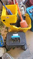 Yellow mop bucket with wringer holding orange plunger and blue scoop, small vacuum and black plastic step stool in front