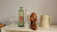 Shelf with decorative items including clear glass decanter, green painted bottle, white ceramic figurine, wooden carved figure, ceramic beer mug.