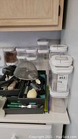 Clear plastic storage canisters stacked and arranged, with various kitchen utensils and flatware grouped in trays underneath. Includes mesh strainer and sharpener.