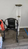 Walker with seat and backrest alongside a pair of aluminum crutches with beige grips and tips on wooden floor.