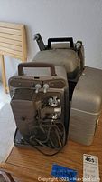 Bell and Howell and Ambico projectors sitting on a wooden table with their cords visible, with Bell and Howell projector front around the lens area and side with controls shown.