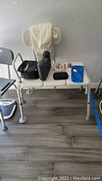 Photo showing beige shower bench with white towel on backrest, black carrying case, black portable scale, and small blue item on bench.