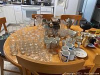 Full view of the lot arranged on the table including glassware, mugs, pitchers, vase, and metal items.