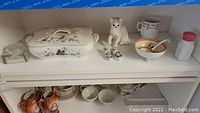 Shelf with covered dish, glass jar, cat figurine, decorative shoes, small bowl with spoon, and pink-lidded container.