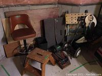 Wide view of vintage barstool, wooden step stool, foam pads, black furniture dolly and accessories against basement wall
