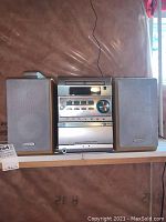 Central Kenwood micro hi-fi stereo system with speakers on each side sitting on a shelf in a basement with insulation visible behind.