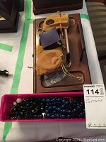 Top-down photo showing a wooden tray holding a gold-tone pocket watch with chain, metal rings, and small pouches. Below the tray are multiple strands of blue and black glass bead necklaces in a purple container.