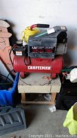 Front view of Craftsman air compressor showing red tank with white Craftsman label, motor housing, and yellow coiled air hose on top.