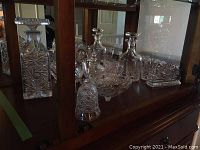 Multiple views of crystal and cut glass barware displayed in cabinet, showing decanters, glasses, swan dish, napkin holder, bell, bowl, and creamer.