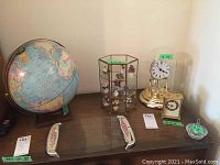 Desk globe, anniversary clock, brass carriage clock, glass case with miniature porcelain flowers, and crystal ring holder arranged on a wooden table.