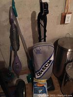 Oreck upright vacuum cleaner with blue and grey bag, vacuum bags included, next to a green mop bucket and cleaning tools.