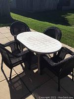 Photo showing the entire patio set with four faux wicker chairs around an oval white resin table on a tiled patio outdoors.