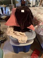 Stack of pillows, linens, and blankets on top of two storage totes, one blue and one gray. Lot items are shown in an indoor basement setting.