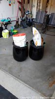 Two black clay planters with buckets stacked inside, one containing potting soil bag, on garage floor.
