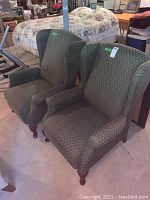 Two matching green patterned upholstered recliners shown side-by-side on a carpeted floor with a bed and other furniture in the background.
