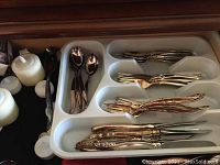 Drawer open showing multiple pieces of silver-plated flatware in a white plastic flatware organizer tray including spoons, forks, and decorative knives with ornate handles.