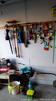 Garage wall showing assorted yard tools hanging on wooden pegboard including umbrellas, gloves, various hand tools, and Disney-themed yard decorations featuring Mickey Mouse and Donald Duck. Bench below holds Ryobi trimmer, white bucket, and some gardening supplies.