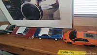 Four model cars in a row on a wooden surface beneath framed car photo