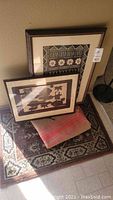 Photo showing two framed vatuue tapa cloth artworks leaning against a wall on top of two small area rugs with visible patterns and colors.