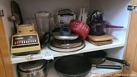 Photo of shelf showing Osterizer blender and base, pots with lids, pans, and cutting boards