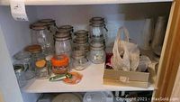 Glass jars of various sizes with orange gasket seals and metal clasps displayed on a shelf, alongside some plastic containers and miscellaneous kitchen items.