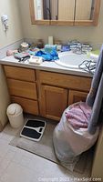 Photo of bathroom vanity area with multiple items including scale, trash can, towels, and various bathroom accessories on countertop under mirror.