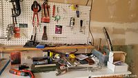 Wide view of various tools and hardware on pegboard, shelf and workbench, including clamps, pliers, screwdrivers, drill bits, screws, and small boxes.