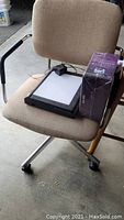 Rolling chair with beige fabric upholstery, light box, power adapter, and boxed item on seat