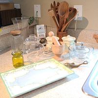Entire lot arranged on kitchen countertop showing wooden utensils in container, ceramic jars, glass teapot, tray, brass bottle opener, and small jars.