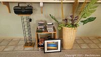 Overall lot view showing wicker basket with artificial plants, stacked CDs, framed photo of canoes, Insignia radio, and small wooden end table with wheels