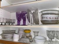 Overall view of shelves holding various kitchen glassware and serveware including Apilco porcelain pie plates, purple wine glasses, clear glass bowls and ramekins.