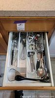 Photo of a kitchen drawer with an assortment of metal and plastic kitchen tools including spatulas, whisks, spoons, tongs and glass scoops organized in plastic tray dividers.