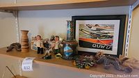 Shelf displaying multiple small handcrafted decor items including figurines, decorated ceramic bottle, pinecone, and framed photo.